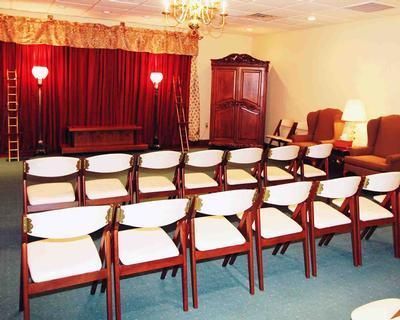 A funeral home chapel with rows of chairs facing a red curtained stage, and a wooden wardrobe.