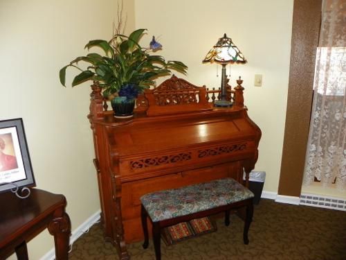 Wooden organ with plant, lamp, and bench in a room with a framed picture and lace curtain.