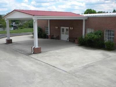 Red-roofed carport entrance to brick building; concrete parking area, small windows, bushes.
