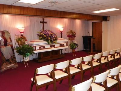 Funeral home interior: casket on a stand with floral arrangement, rows of chairs, wooden cross.
