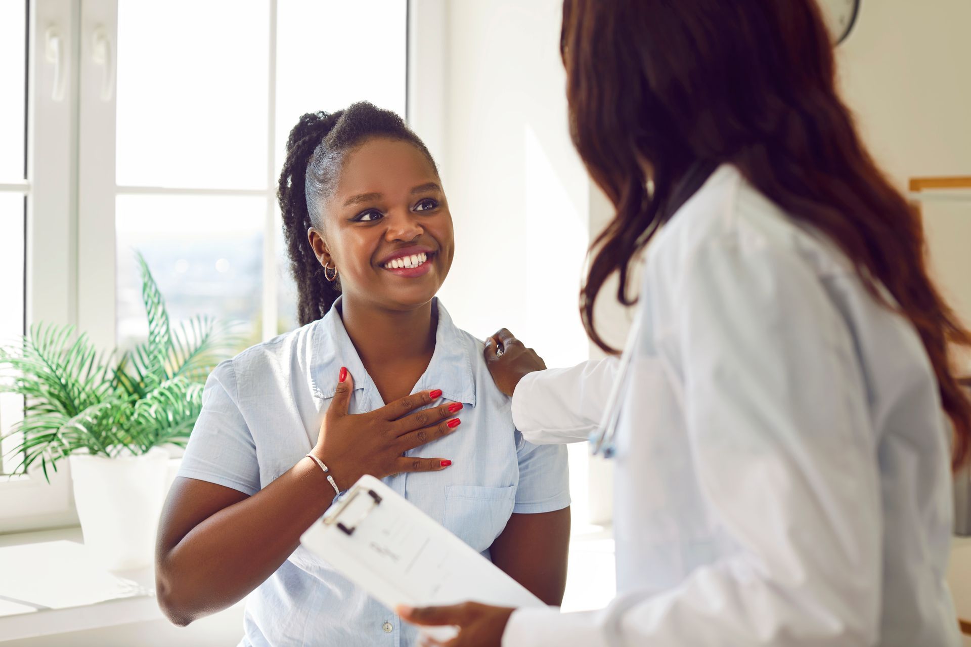 Black woman smiles at doctor, hand on her chest. Doctor has hand on her shoulder. Indoors.