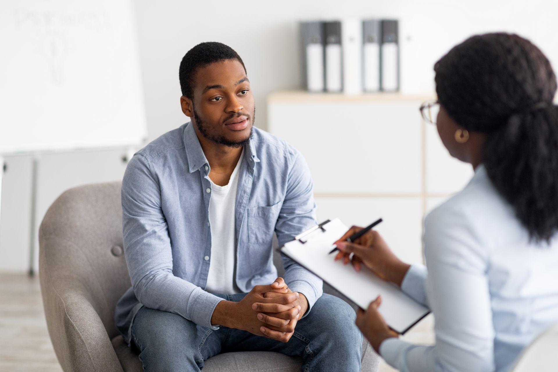 Man in a therapy session, seated, speaking to a woman taking notes in a light, modern office.