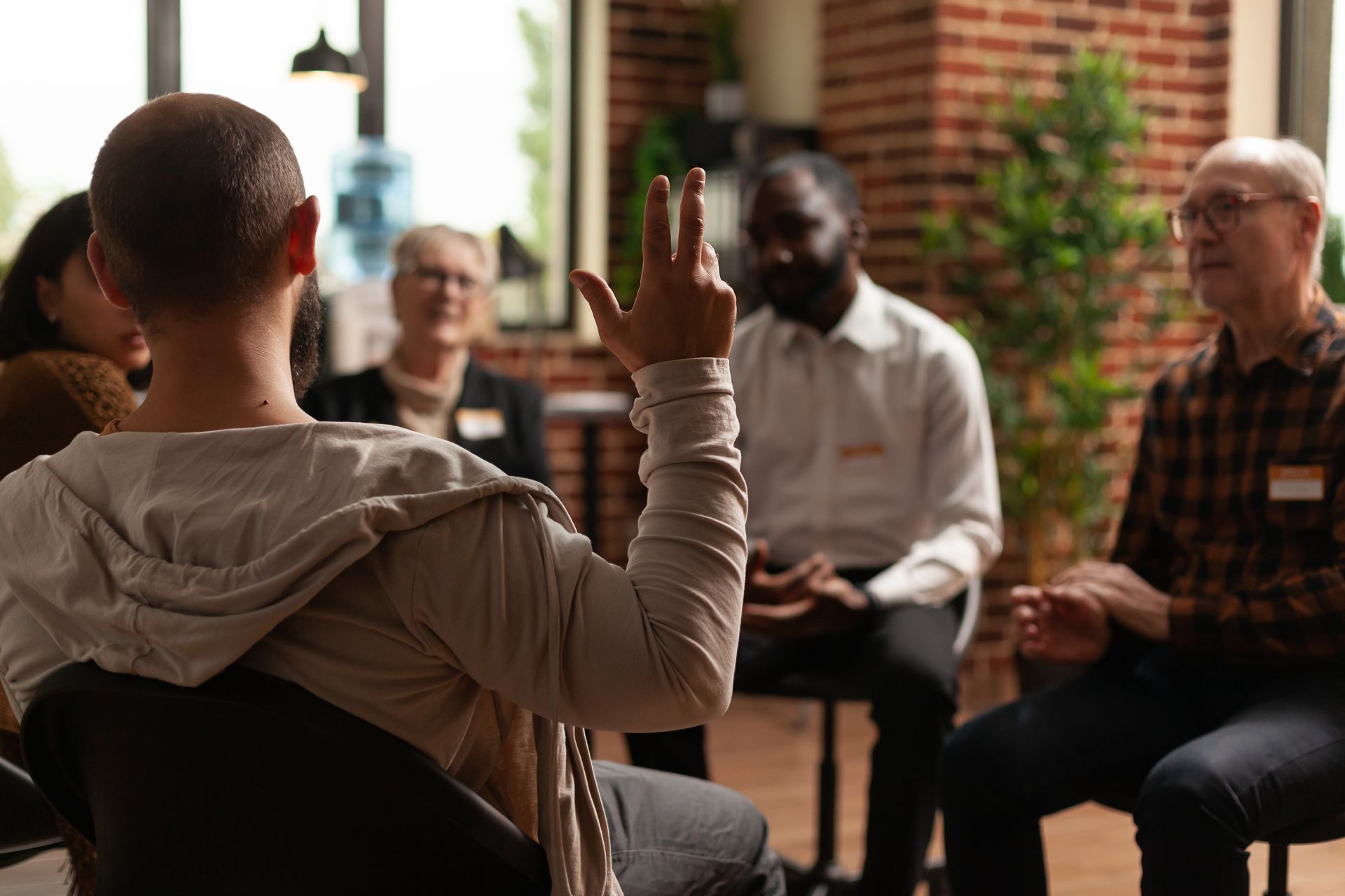 Group therapy session, man raising hand to speak; diverse group in a circle indoors.