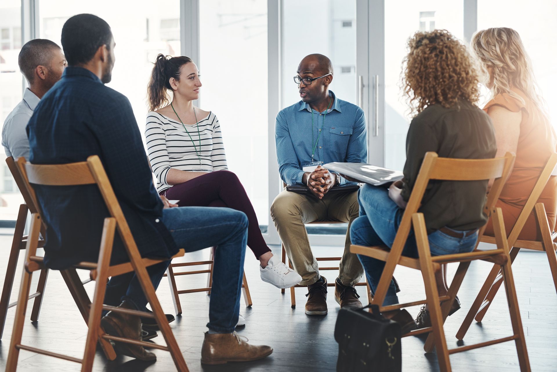 Group therapy session: People seated in a circle talking, led by a man holding a notepad, in a bright room.