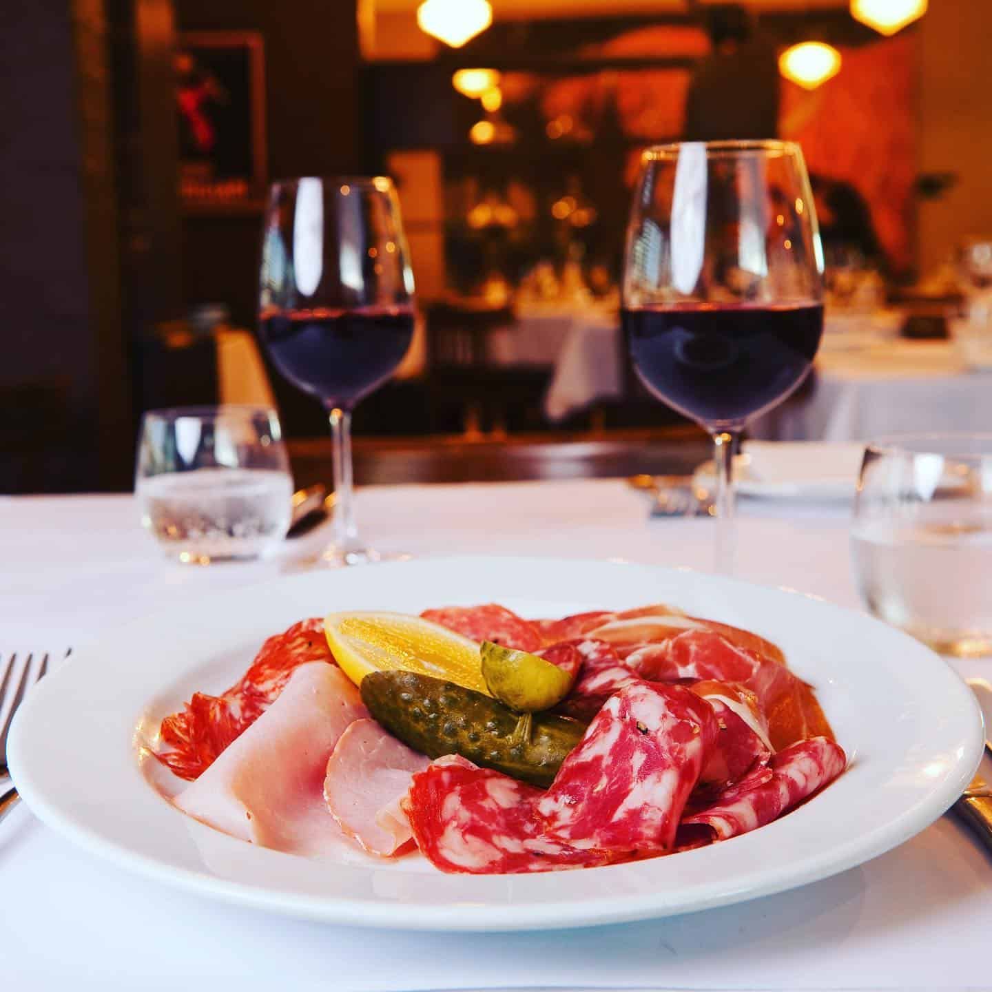 A Plate of Meat and Pickles on a Table With Two Glasses of Wine — Onesta Cucina in Bowral, NSW