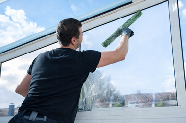 A person in a black shirt uses a squeegee to clean a large glass window outdoors against a blue sky.