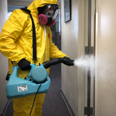 A person in a yellow hazmat suit and respirator sprays a disinfectant mist onto a doorway in a hallway.