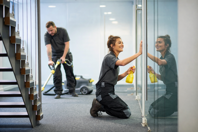 Two professionals in uniforms cleaning an office space; one vacuuming the carpet, the other wiping a glass partition.