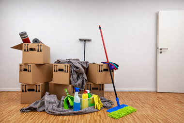 Cardboard boxes, cleaning supplies, and a mop are arranged on a wooden floor in front of a white wall and door.