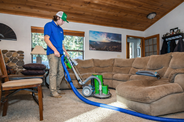 A worker uses a green carpet-cleaning machine on beige carpeting in a living room with a large tan sectional sofa.