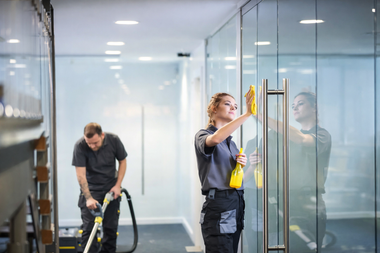 Two professional cleaners in grey uniforms tidy a bright office with a glass partition, using a vacuum and a cloth.