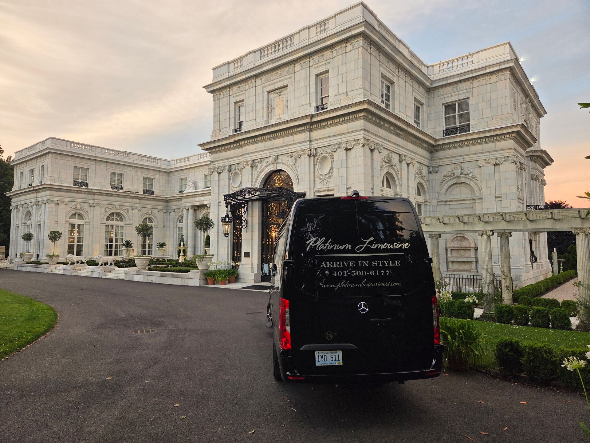 A black luxury transport van parked in front of a grand, white stone mansion at sunset.