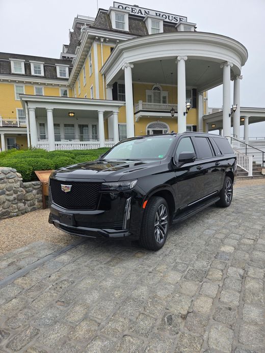 A black Cadillac SUV parked on a cobblestone driveway in front of a yellow historic building with a large porch.