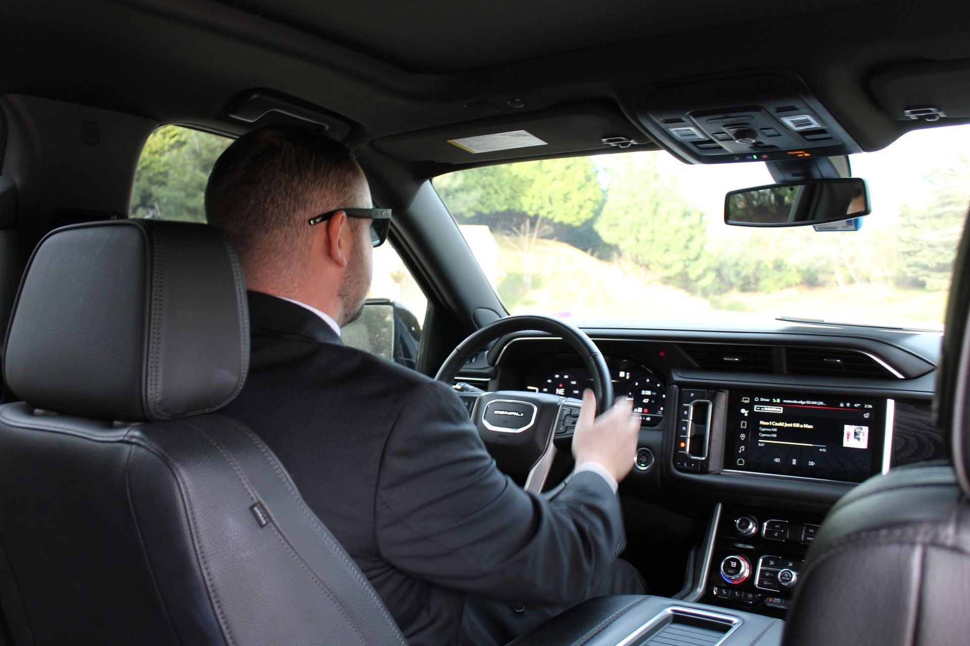 A person in a suit drives a modern vehicle along a road lined with trees.