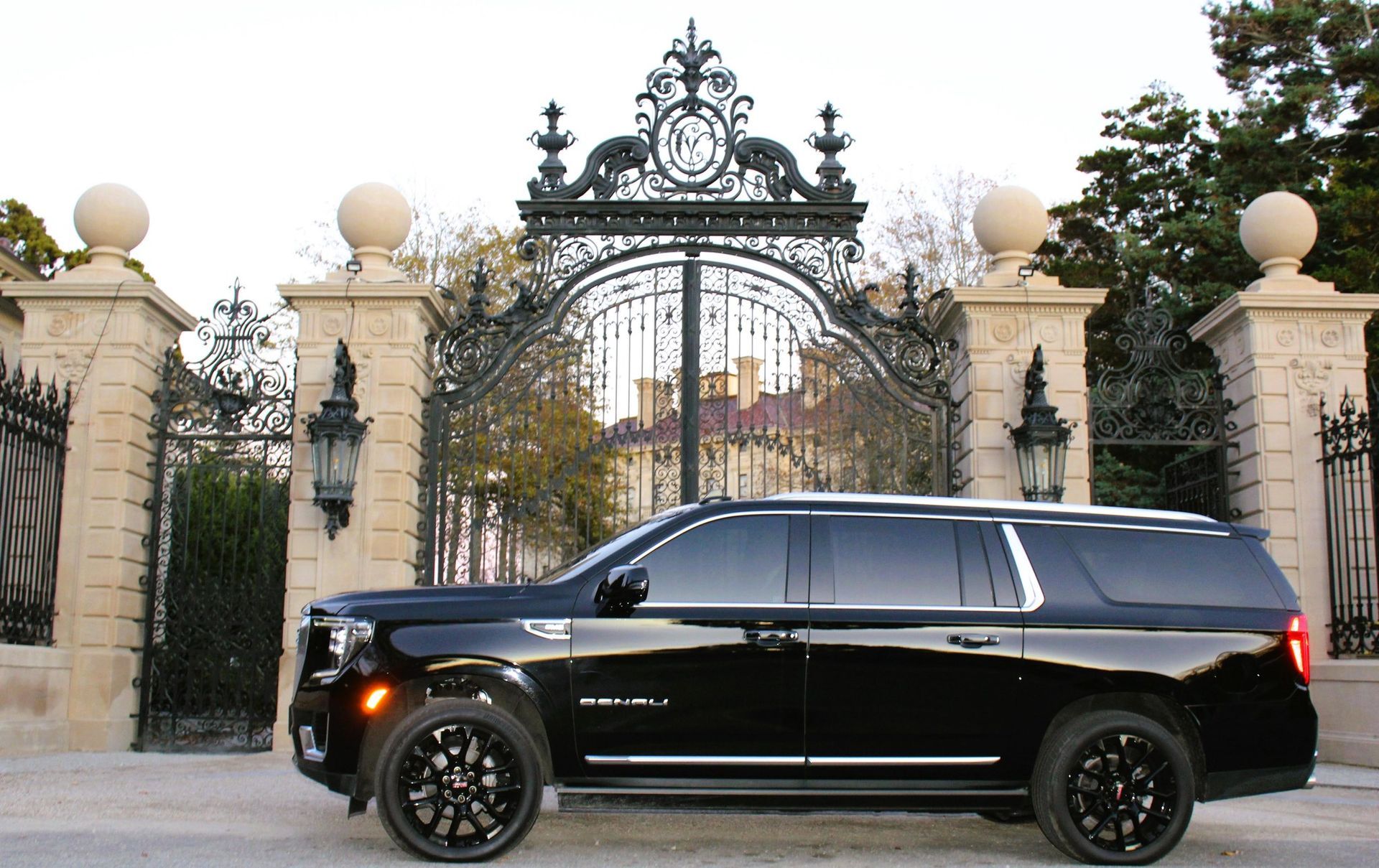 A black GMC Yukon Denali SUV parked in front of a grand, ornate metal gate and stone pillars.
