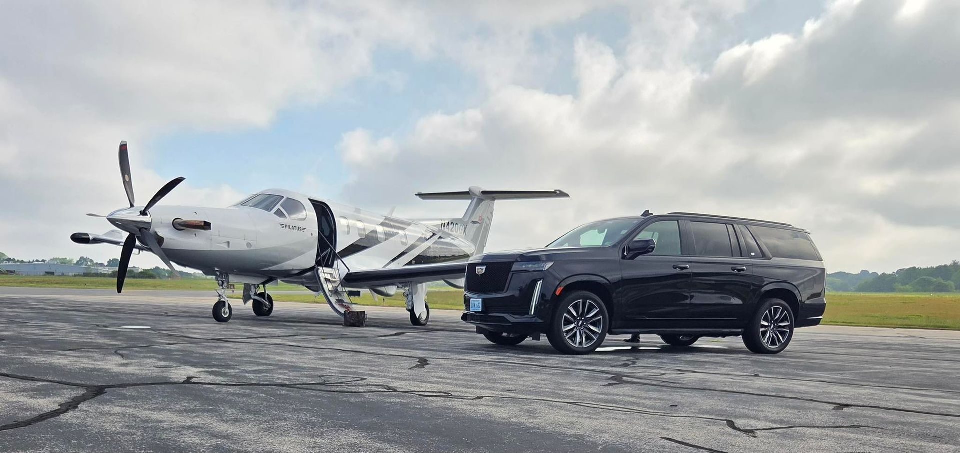 A black Cadillac SUV parked next to a white turboprop airplane on an airport tarmac under a cloudy sky.