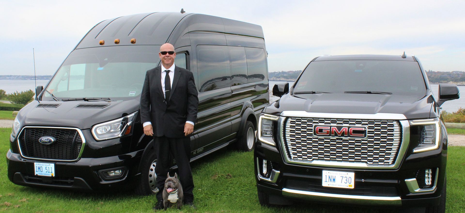 A professional in a suit and a small dog stand between a black Ford passenger van and a black GMC SUV on a grassy field.