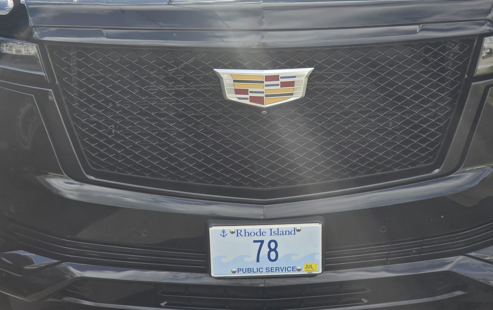A close-up of a black Cadillac grille with a prominent crest emblem and a Rhode Island license plate reading 