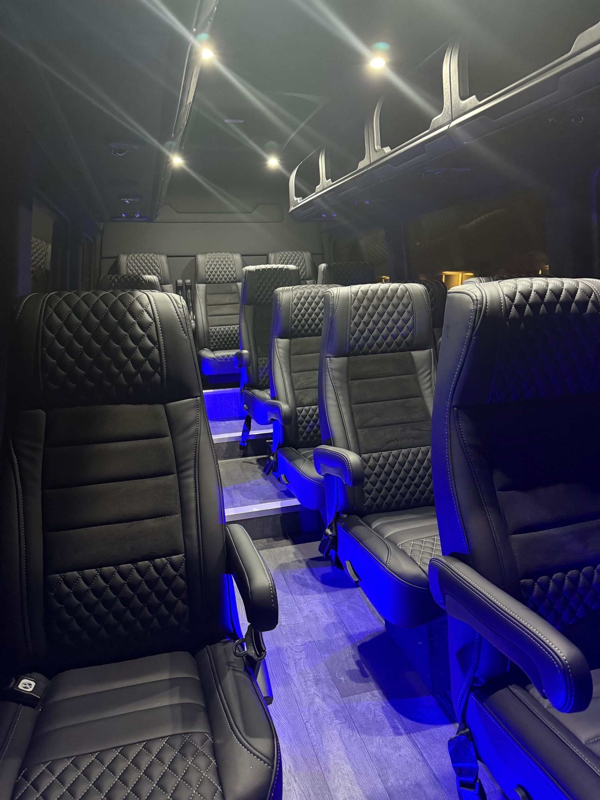 Interior of a luxury passenger van featuring rows of black quilted leather captain's chairs with blue ambient floor lighting.