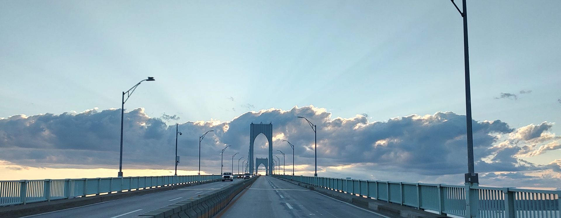 A long bridge stretches toward a large suspension bridge tower under a cloudy sky during sunset.