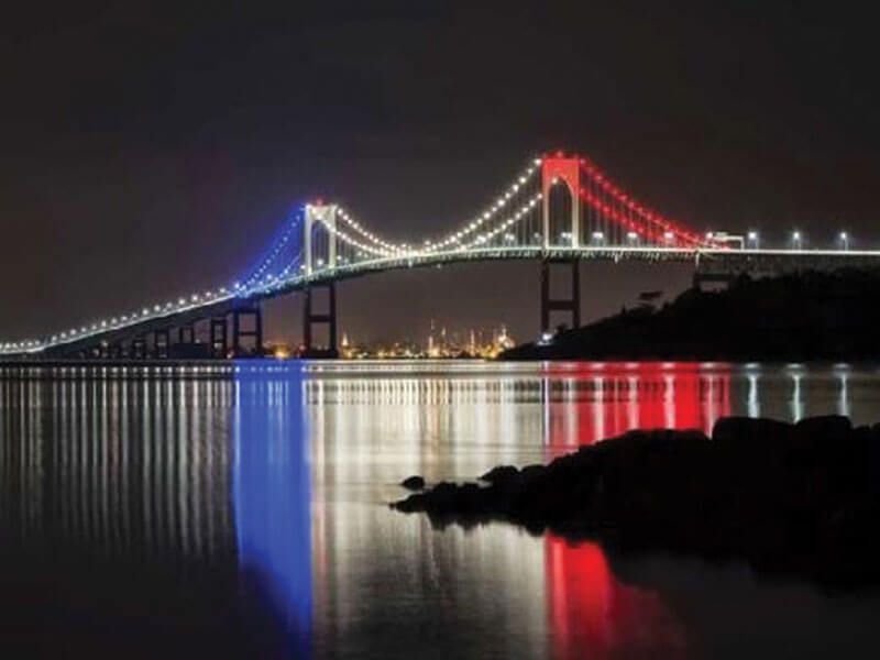 A suspension bridge at night, lit in blue, white, and red, reflecting over calm water.