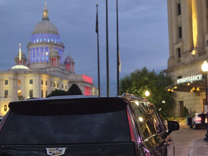 A black Cadillac SUV parked at dusk near the illuminated Wisconsin State Capitol and the Renaissance hotel.