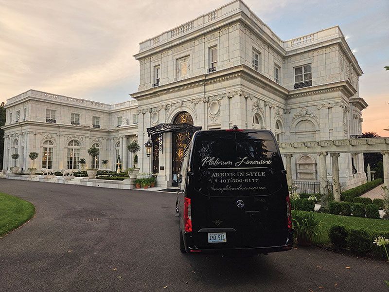A black van parked in front of a grand, white stone mansion at sunset.