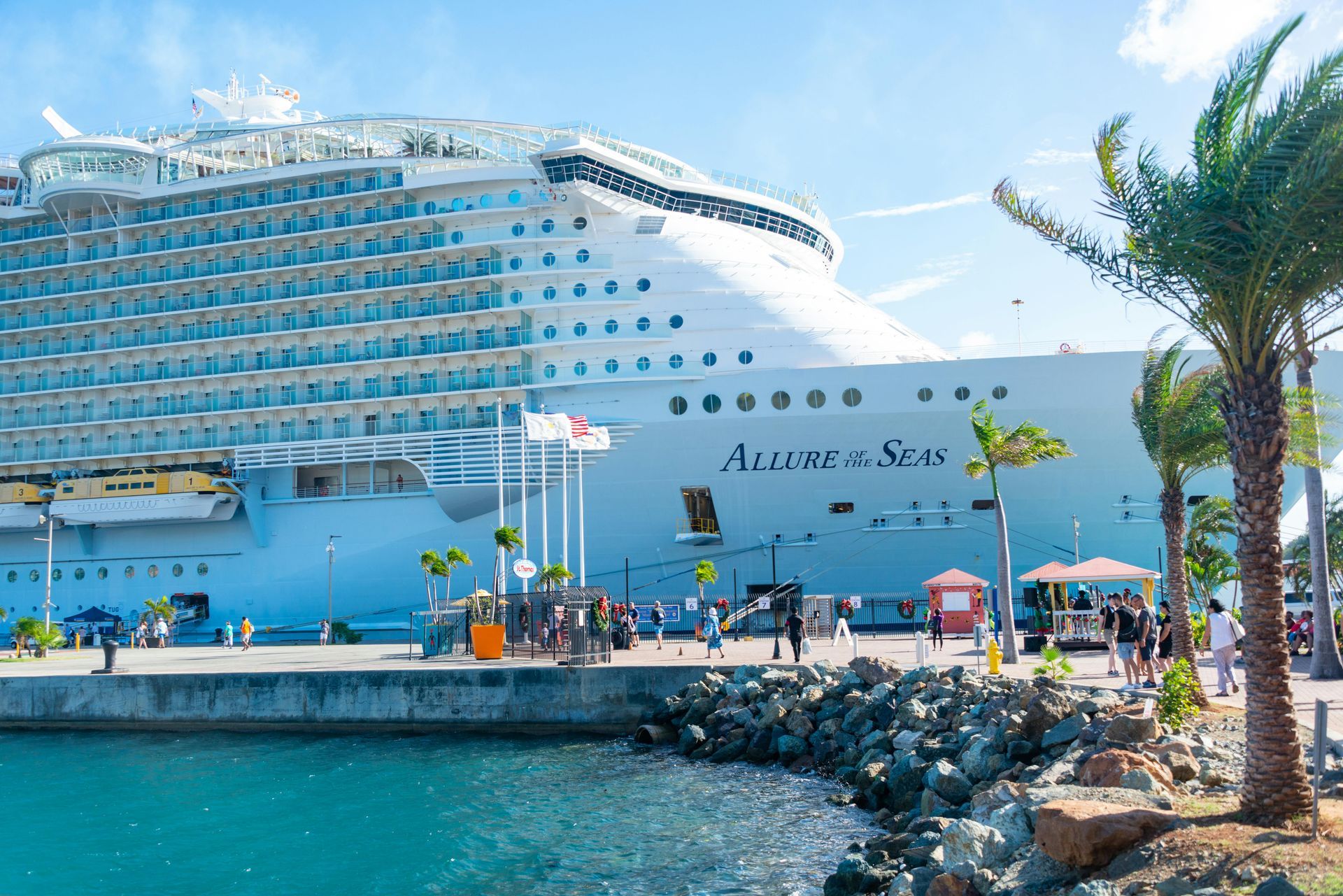 Allure of the Seas cruise ship docked at a sunny port, with palm trees in the foreground and a turquoise sea nearby.