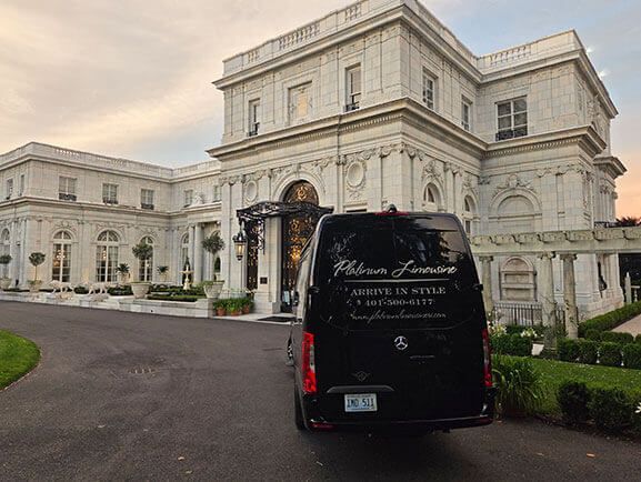 A black luxury passenger van parked in the driveway of a large, white neoclassical mansion at sunset.