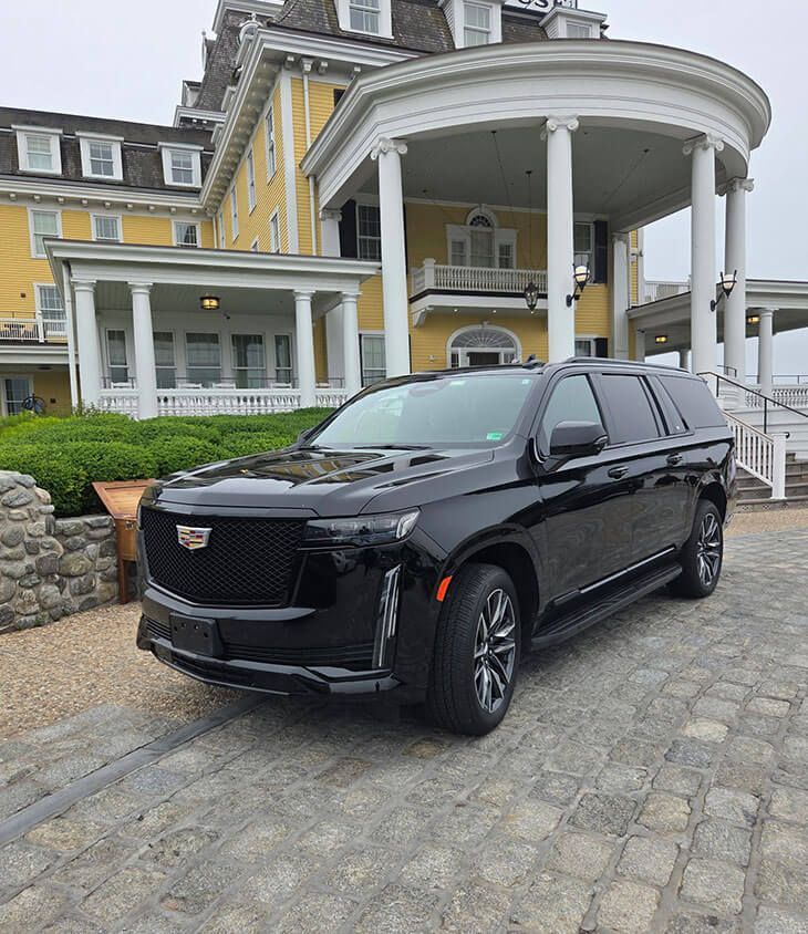 A black Cadillac Escalade parked on a stone driveway in front of a yellow historic mansion with white columns.