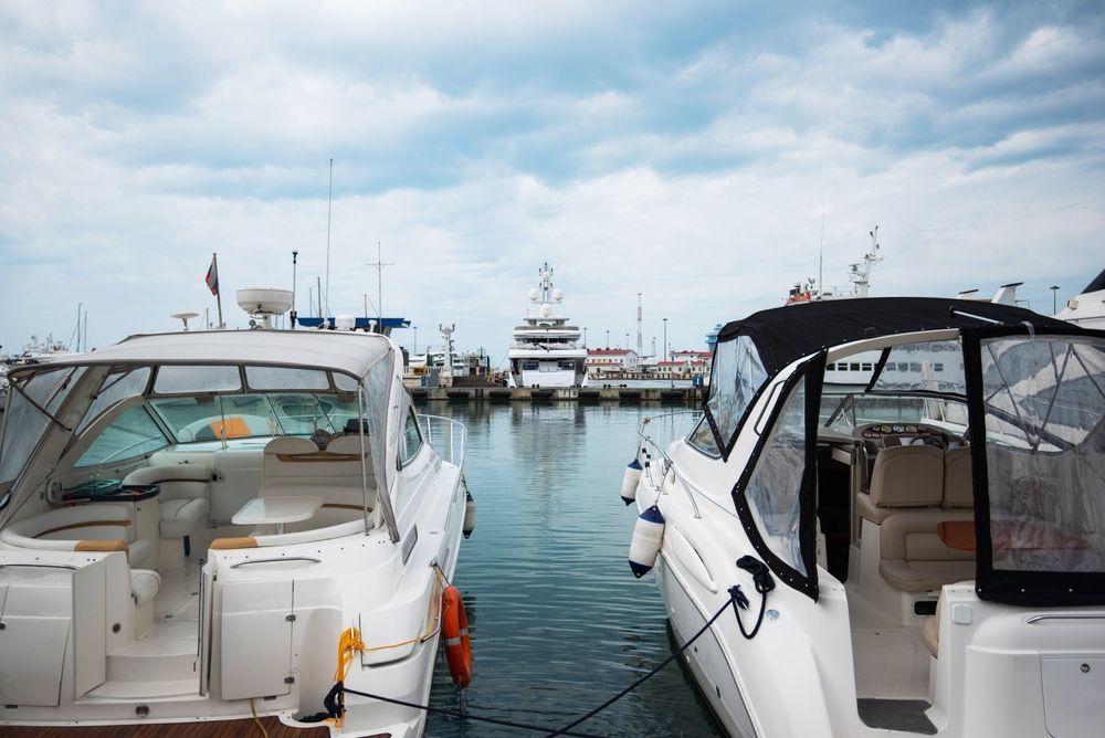Two white motorboats docked side-by-side in a marina, facing a large yacht in the distance under a cloudy sky.
