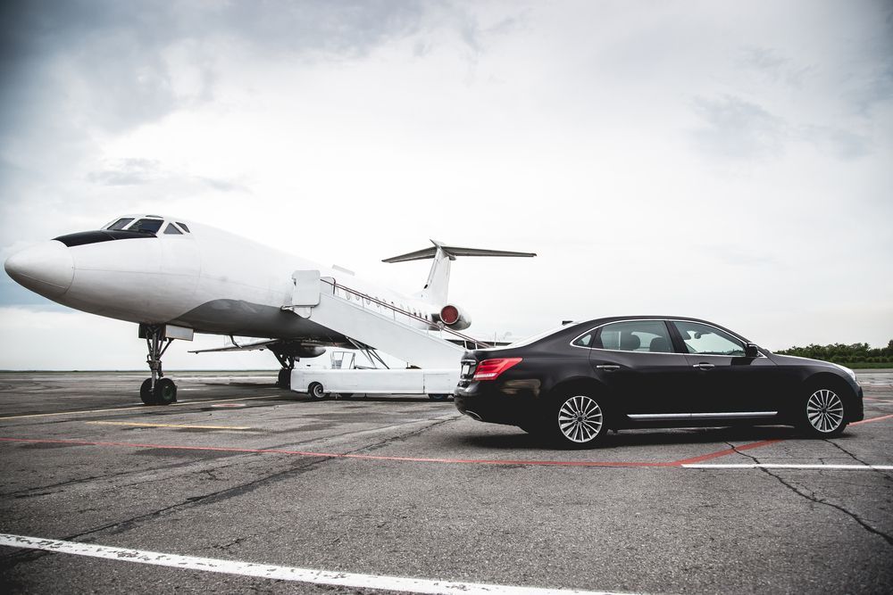 A dark sedan parked on an airport tarmac next to a white private jet with its stairs lowered.