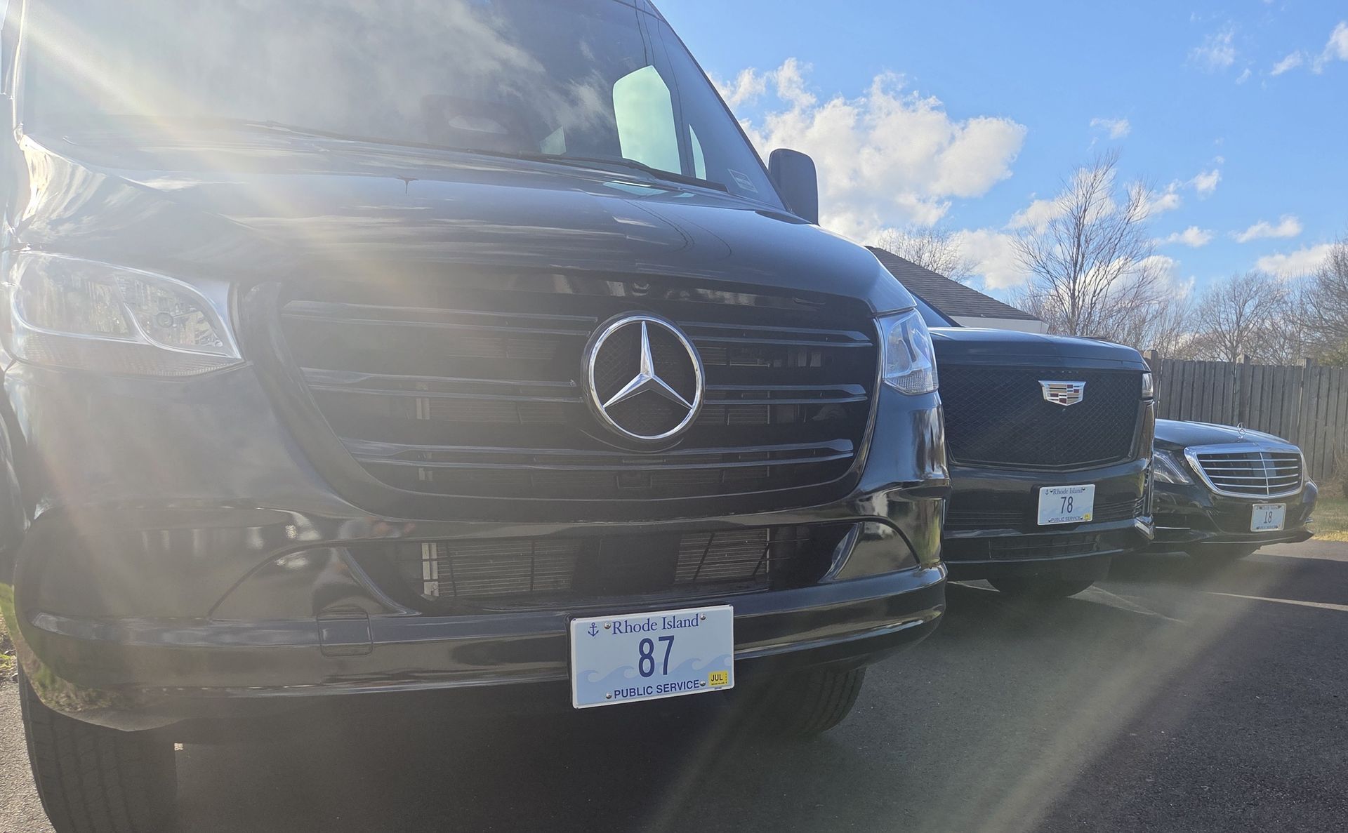 Three black luxury vehicles, including a Mercedes-Benz van, parked in a row outdoors under a sunny blue sky.