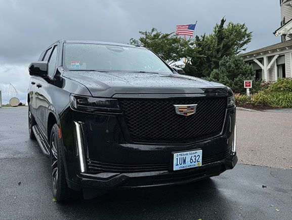 A black Cadillac Escalade SUV parked on a paved driveway with an American flag flying in the background.