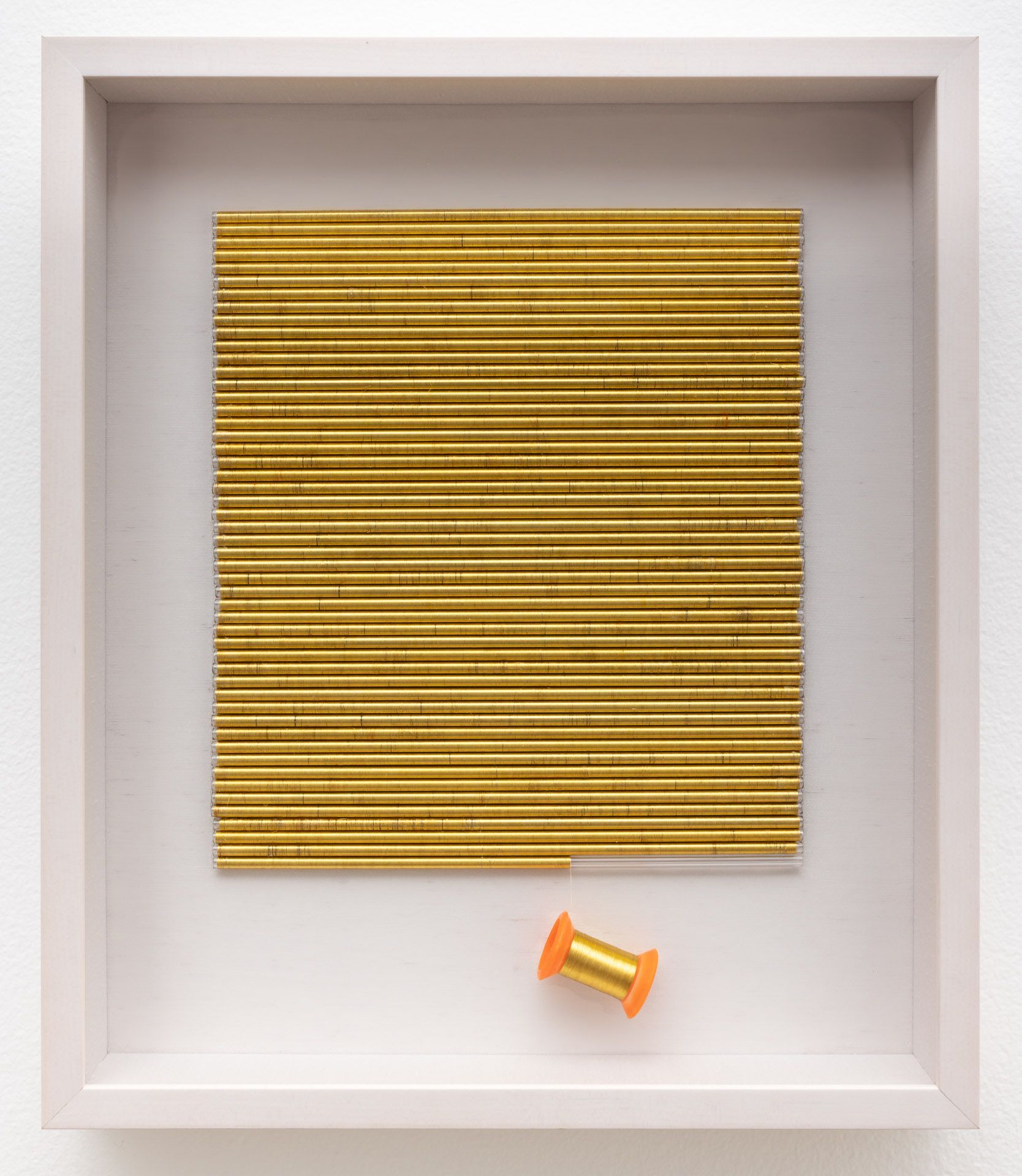 A spool of thread is next to a row of gold sticks in a white frame.