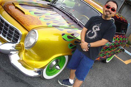 A man is standing in front of a yellow car with flames on it