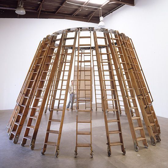 A man stands in front of a dome made of wooden ladders