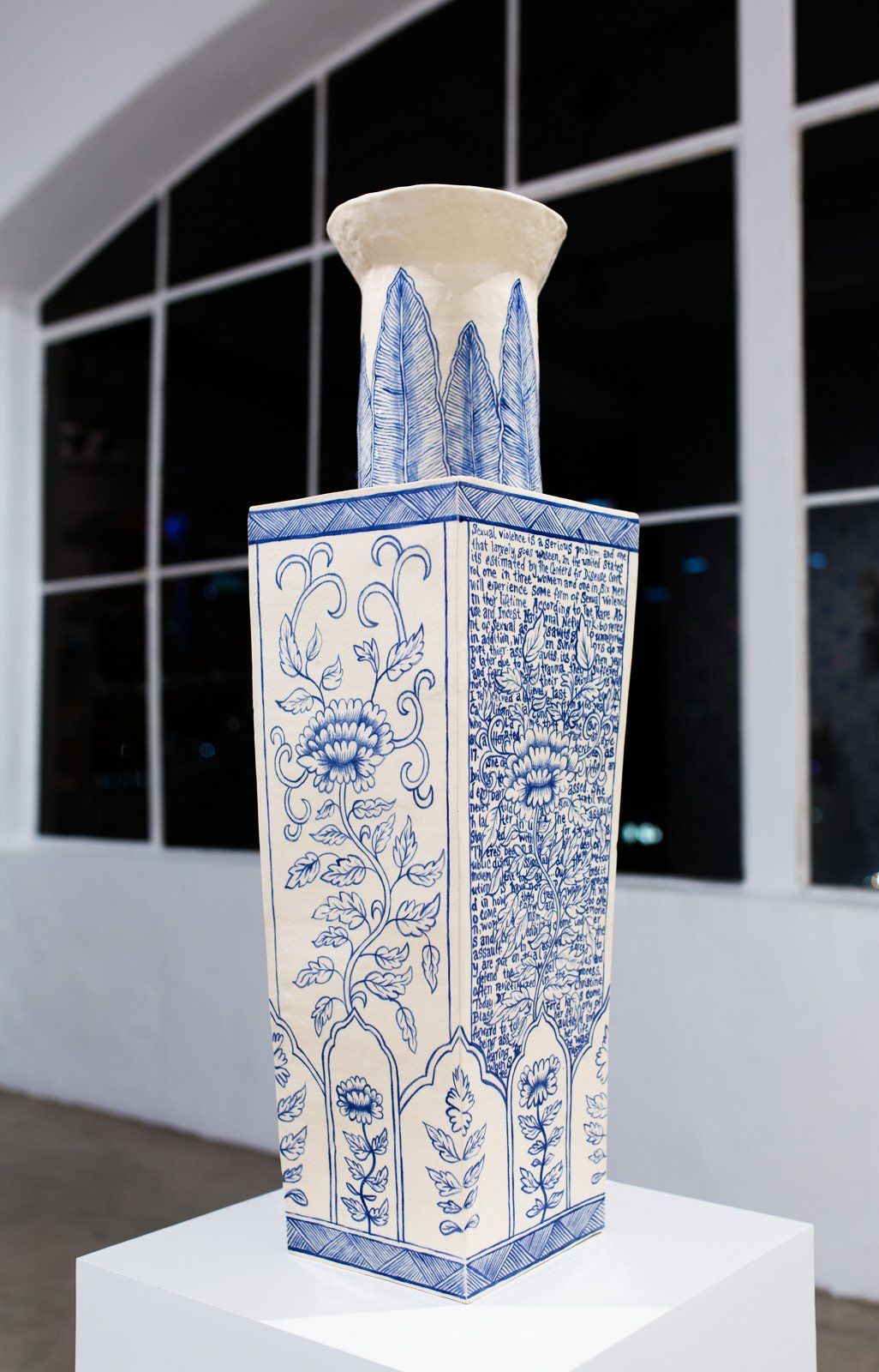 A blue and white vase is sitting on a white pedestal in front of a window.
