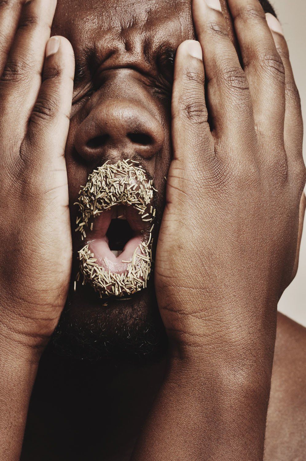 A man with a donut in his mouth is covering his face with his hands