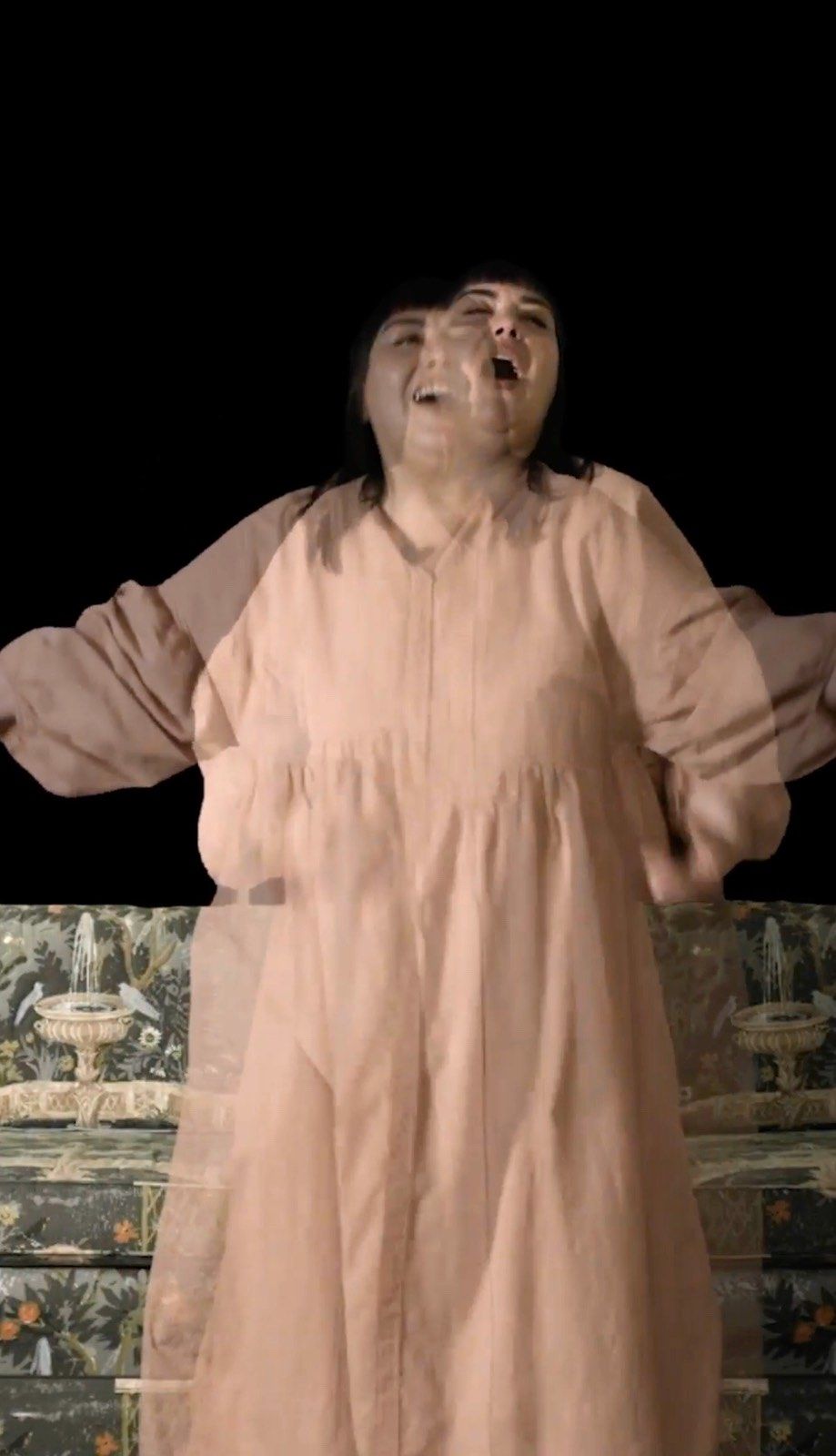 A woman in a pink dress is standing in the dark with her arms outstretched.