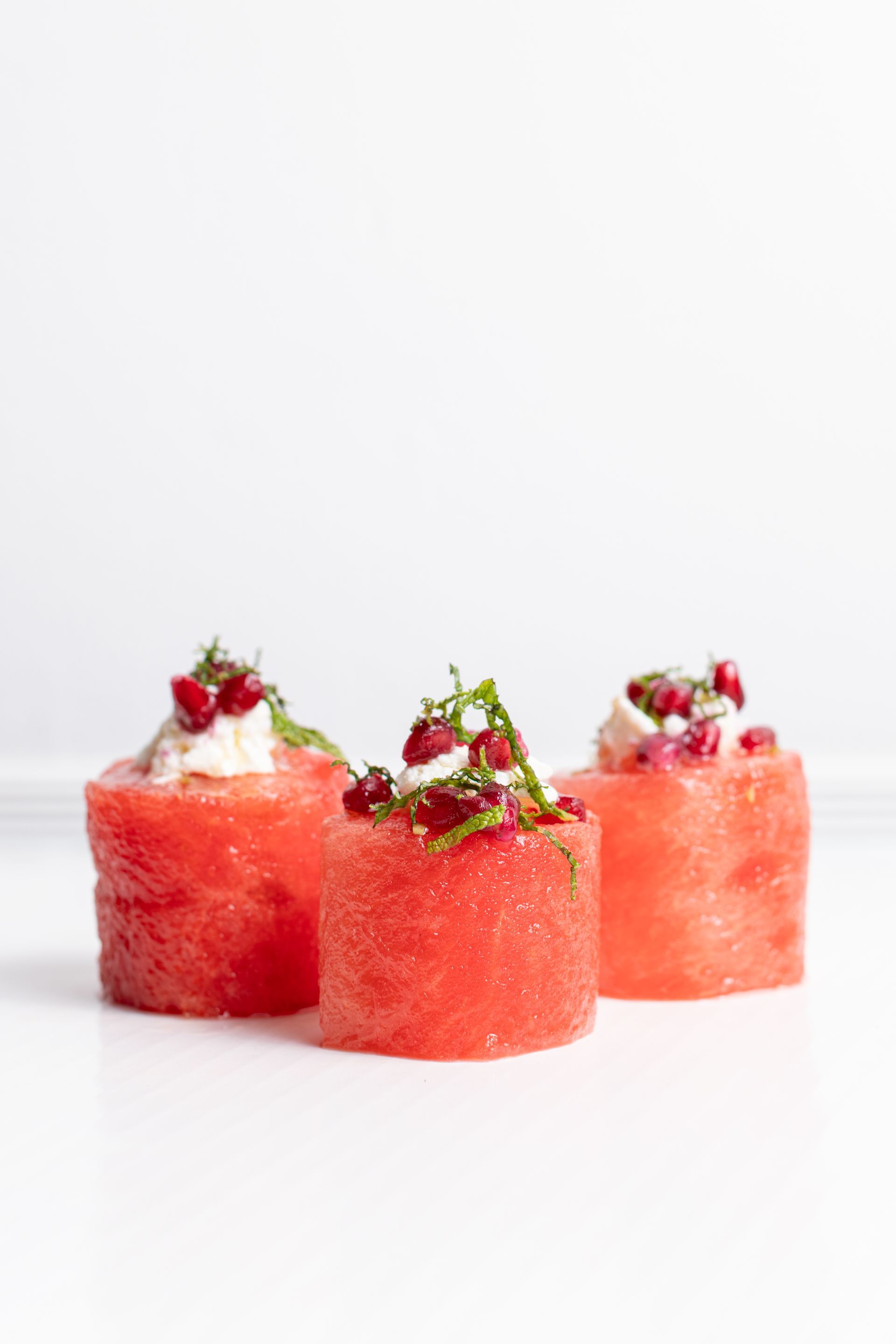Three watermelon slices with feta cheese and pomegranate seeds on a white surface.