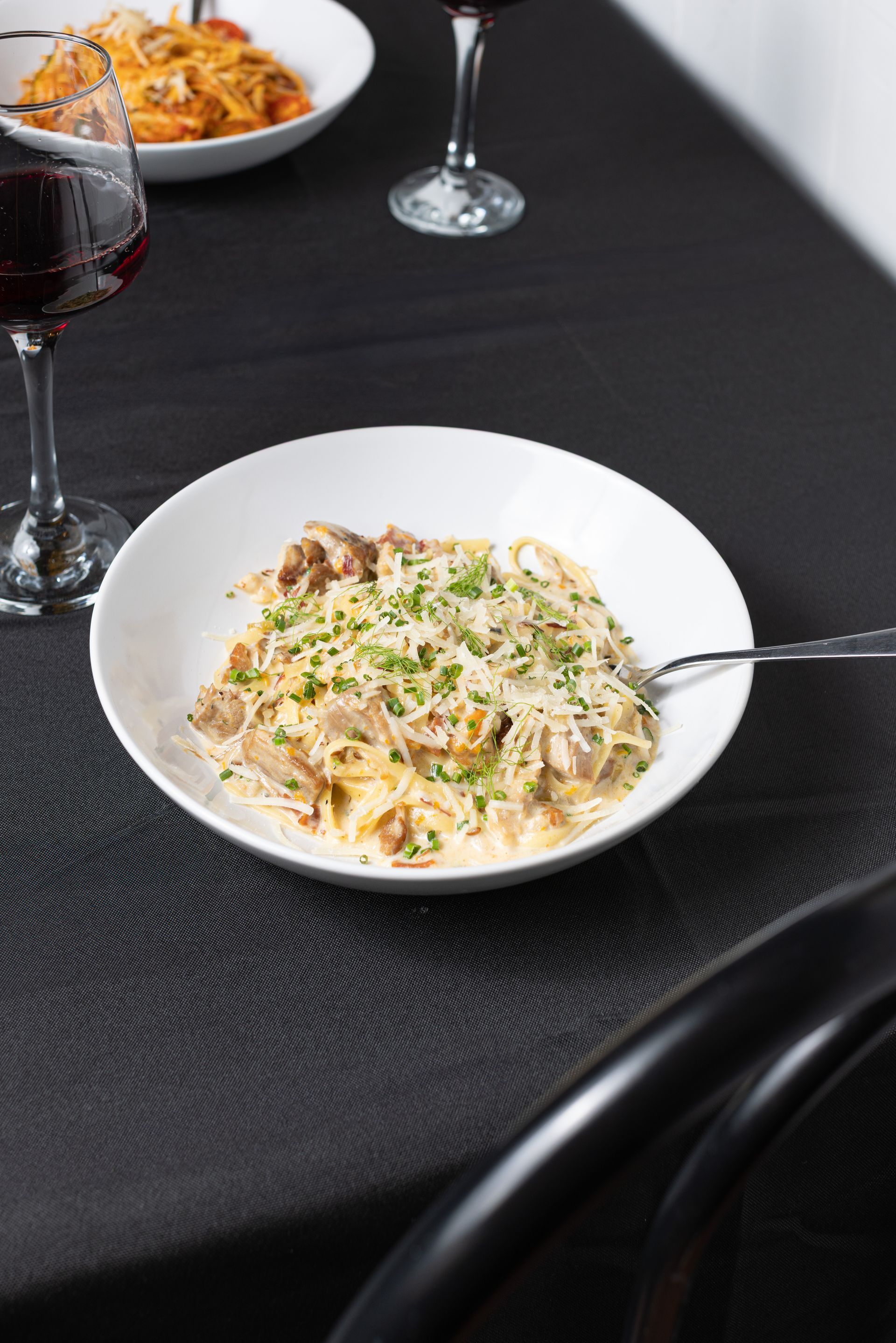 A bowl of pasta with a fork on a table next to a glass of wine.