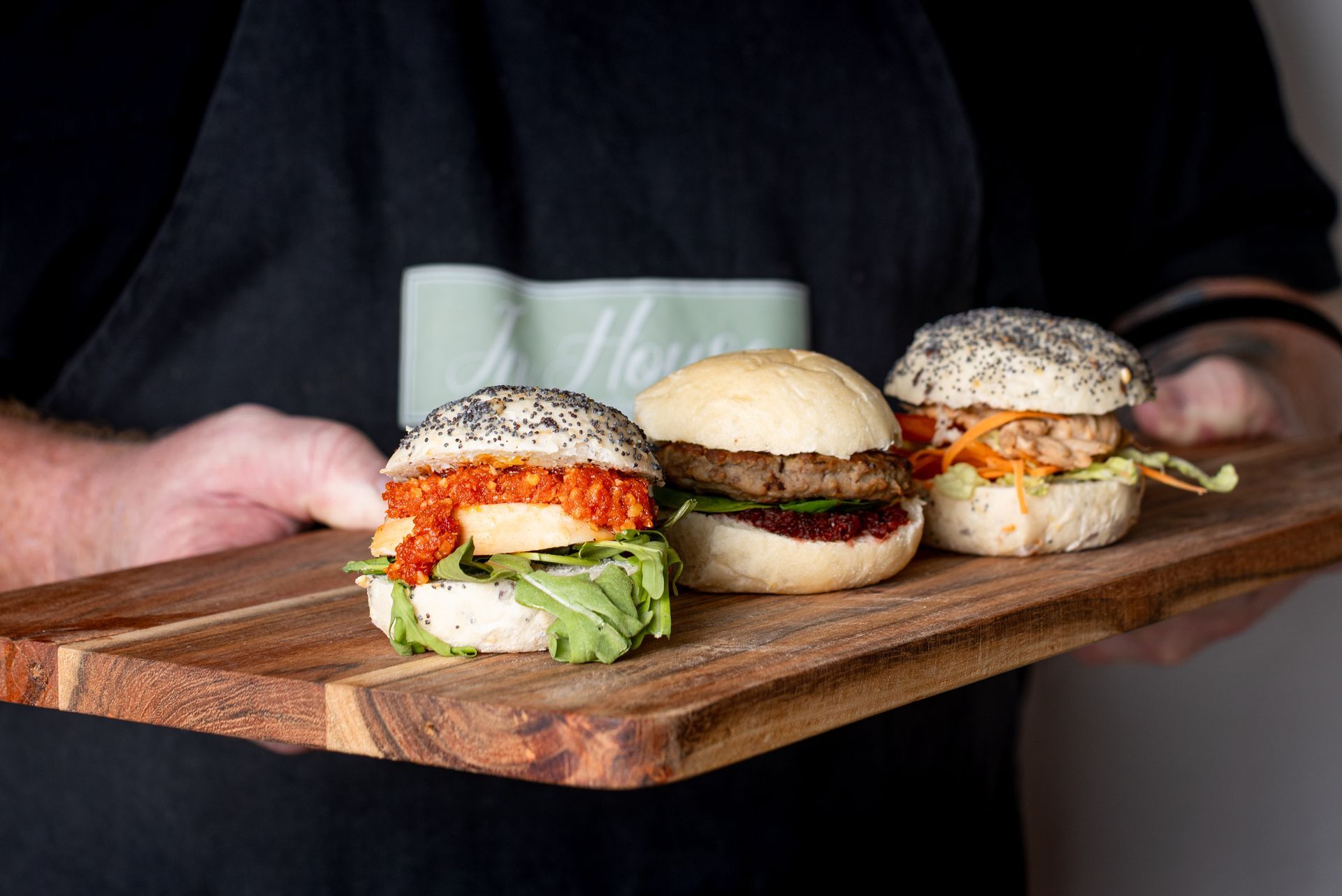 A person is holding a wooden cutting board with three hamburgers on it.