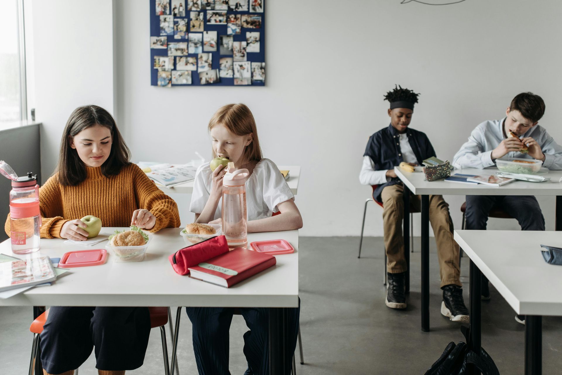 A group of young people are sitting at tables in a classroom eating food.