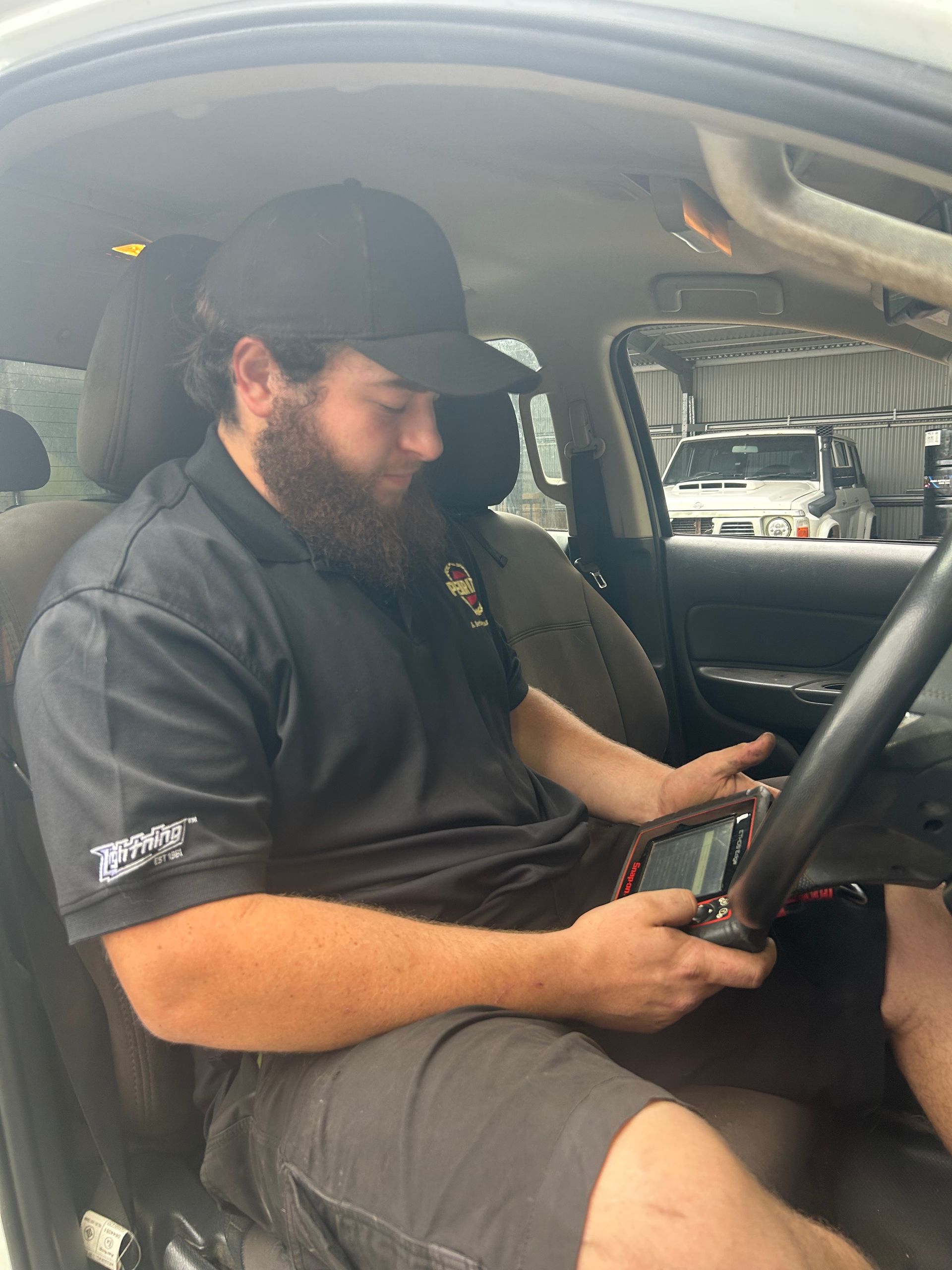 Mechanic in a car using a diagnostic tool. Wearing a black cap and shirt, looking at the device.  — Gympie Diesel & Dyno Centre in Glanmire, QLD