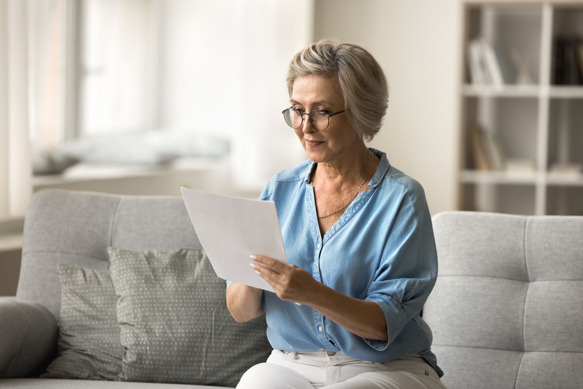 Woman with glasses reading a document while seated on a gray couch in a living room