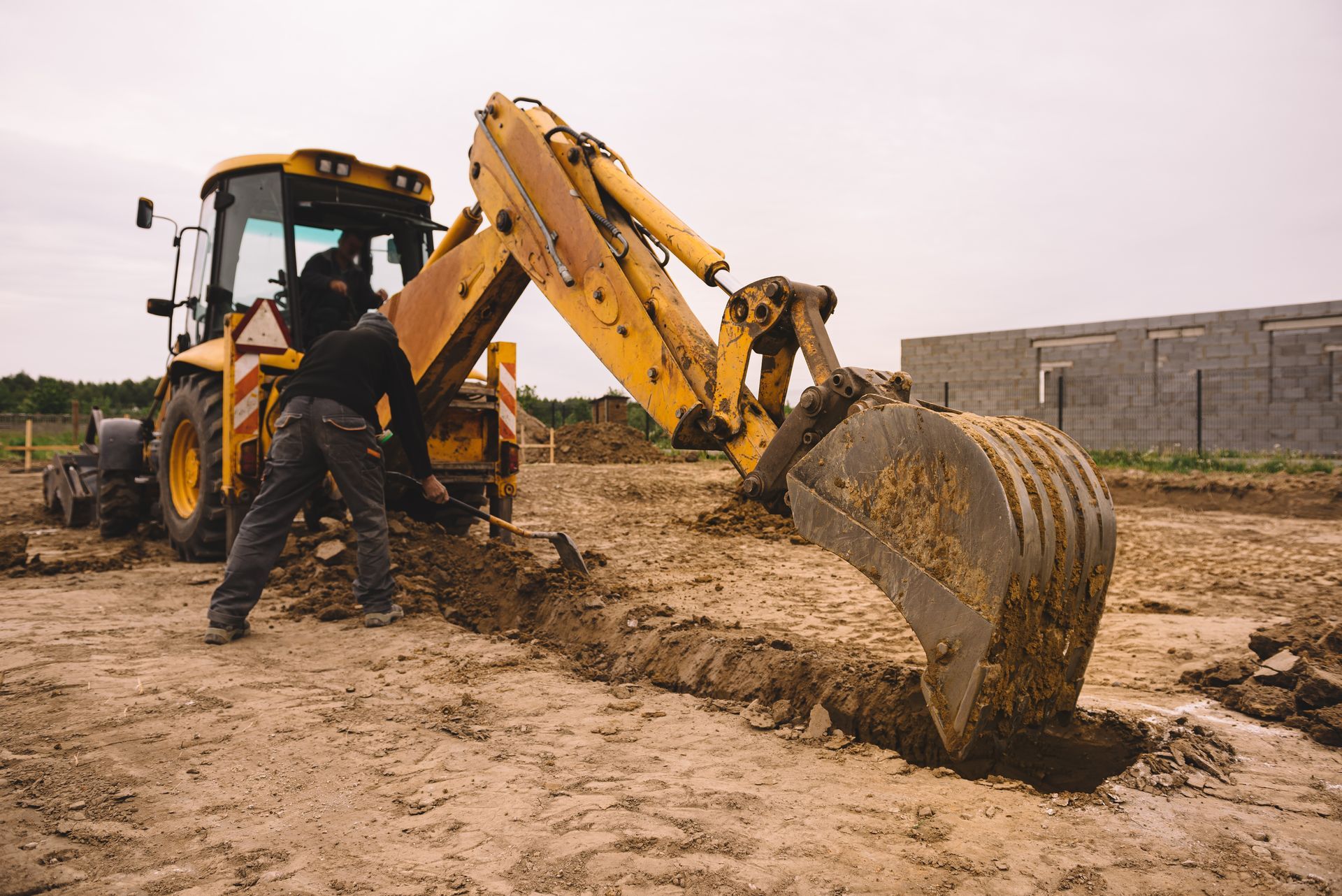 Yellow backhoe digging trench on a dirt construction site; worker nearby.