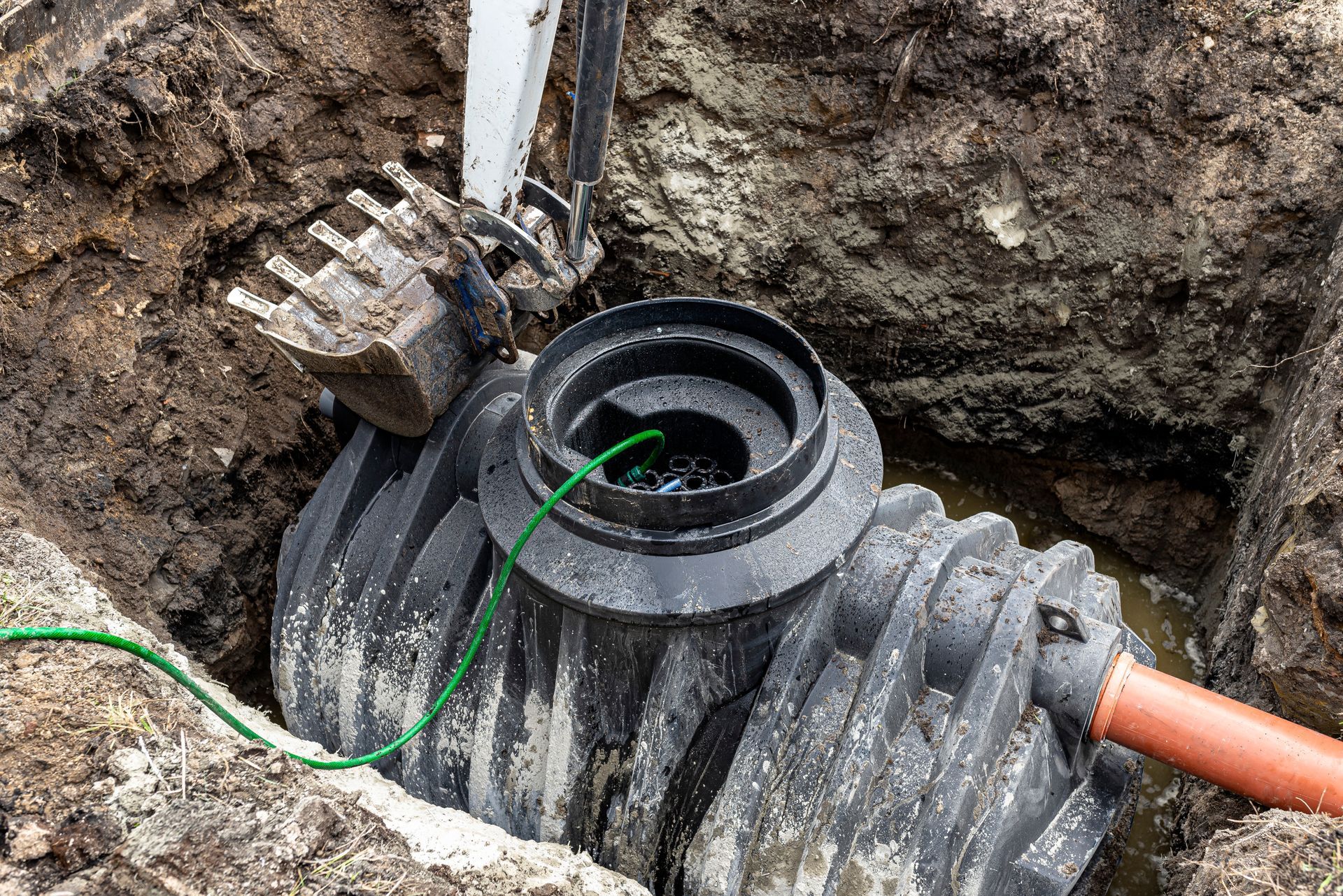 Septic tank installation: Black tank in a dug pit, orange pipe, backhoe bucket overhead.