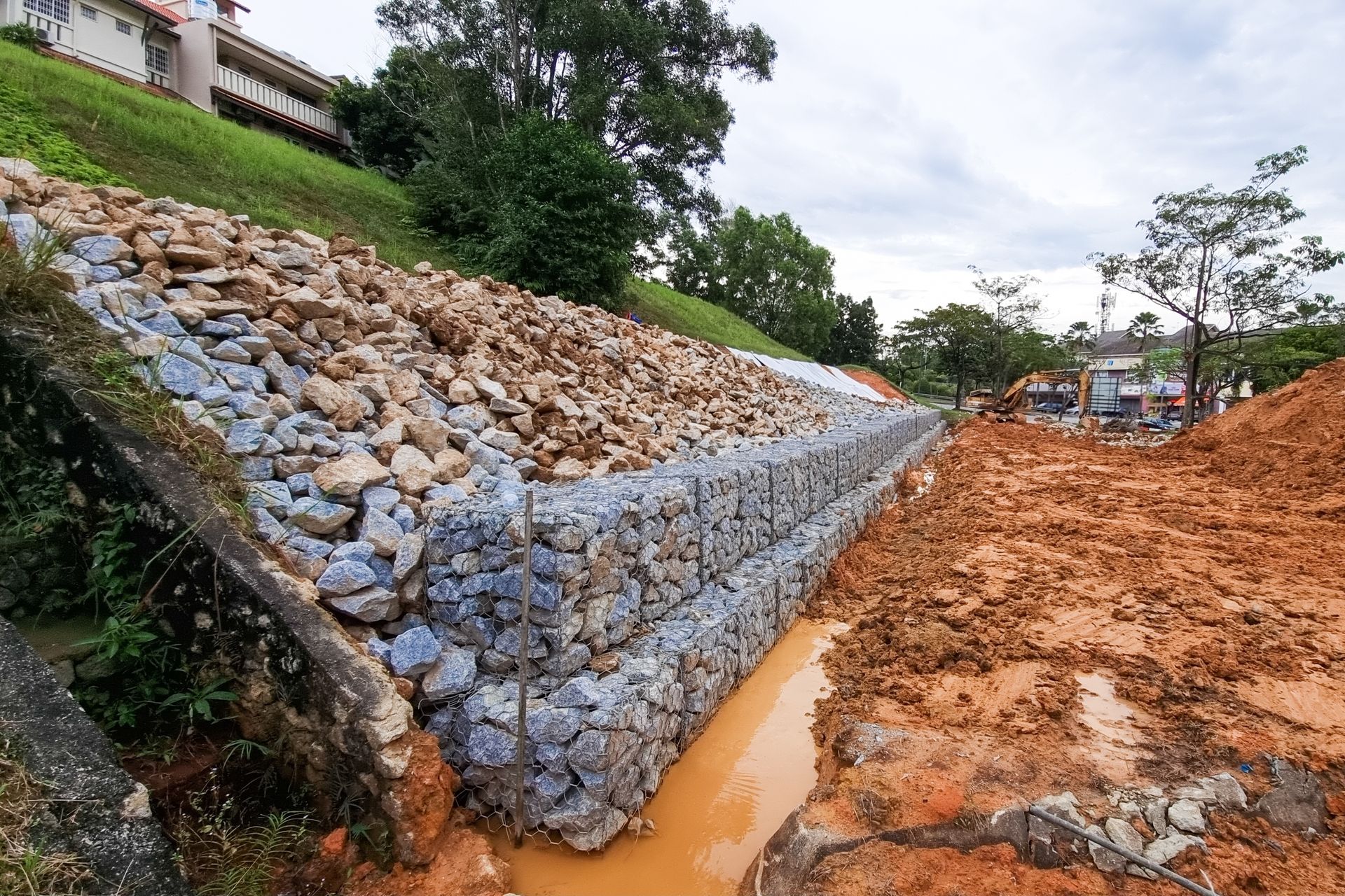 Stone-filled wire cages create a retaining wall on a muddy hillside, adjacent to a dirt construction area.