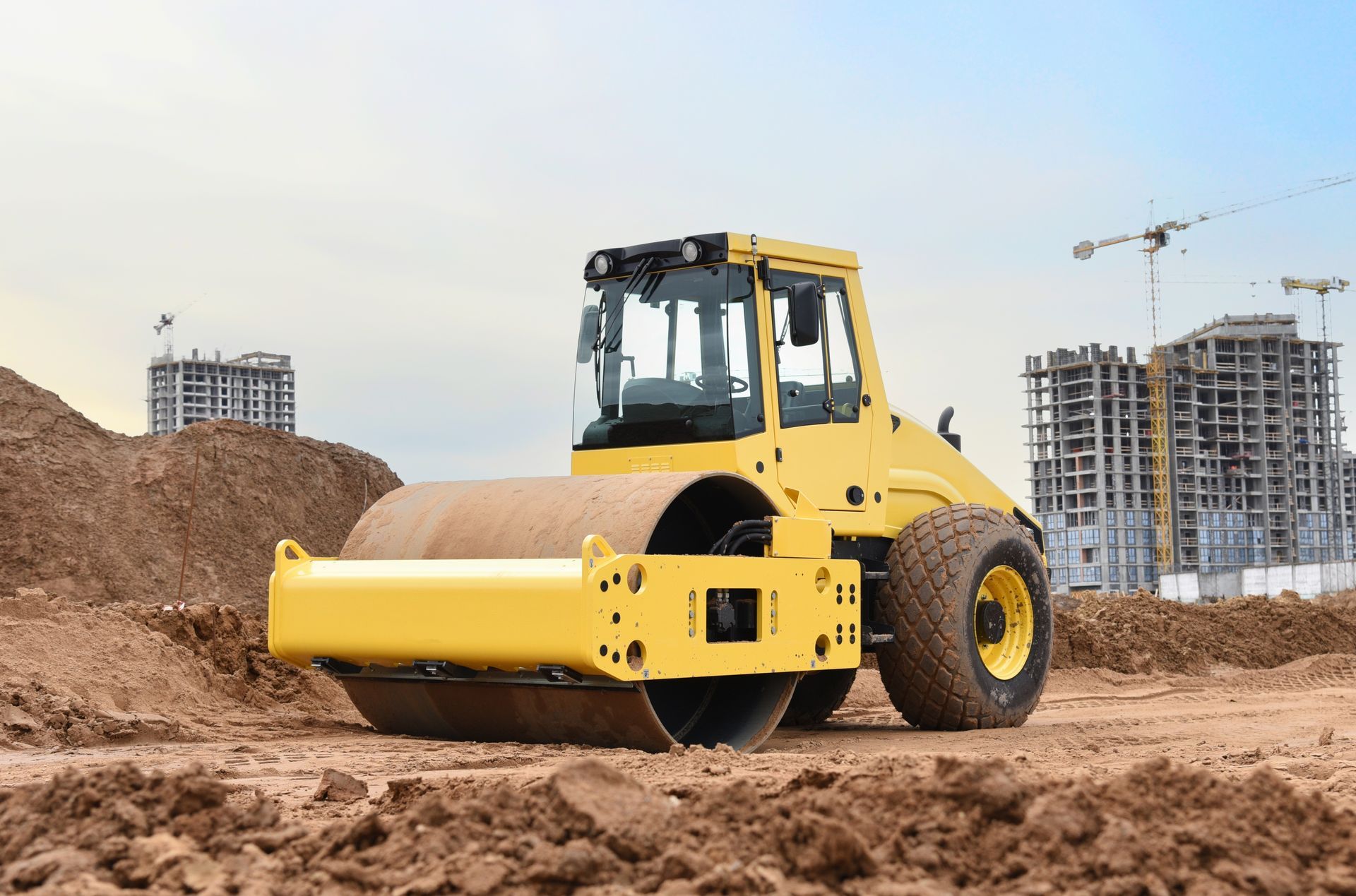 Yellow road roller compacting soil at a construction site, with buildings and cranes in the background.